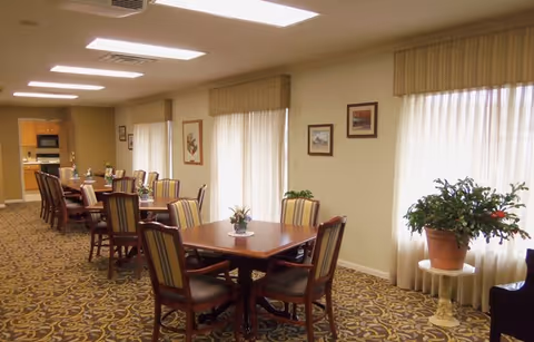 A well-lit dining room with multiple wooden tables and striped cushioned chairs arranged neatly. The room has patterned carpet flooring, large windows with beige curtains, framed artwork on the walls, and a potted plant on a small round table near the window. The far end of the room leads to a kitchen area.