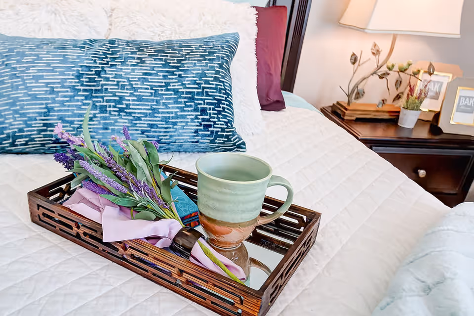 A decorative tray holding a mug and lavender sits on a neatly made bed with pillows and a bedside table and lamp in the background.