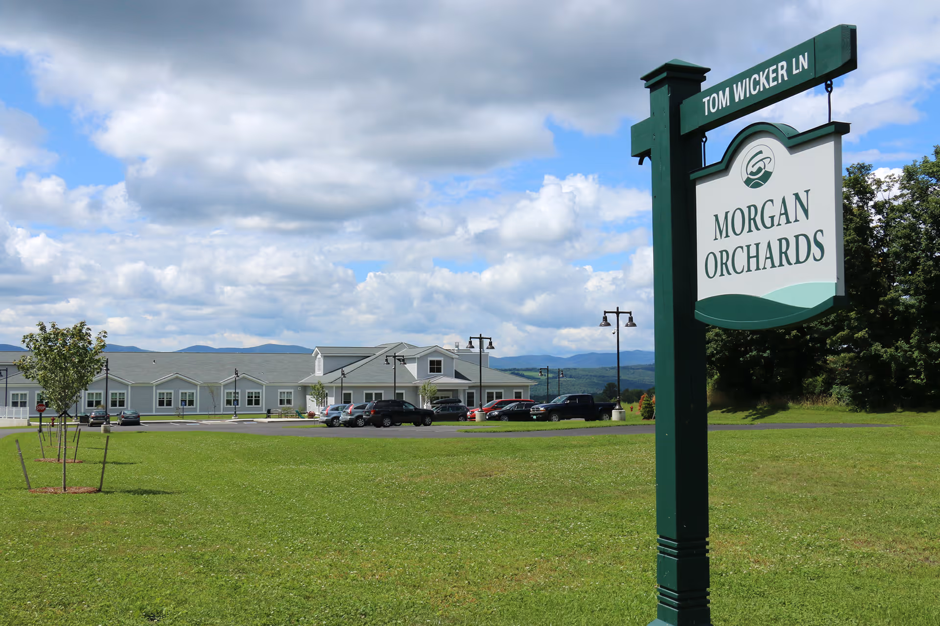 A large green lawn with a street sign labeled 'Tom Wicker Ln' and a hanging sign for 'Morgan Orchards' in the foreground. In the background, there is a long, single-story building with a gray roof and multiple windows, several parked cars, and a partly cloudy sky with distant hills.