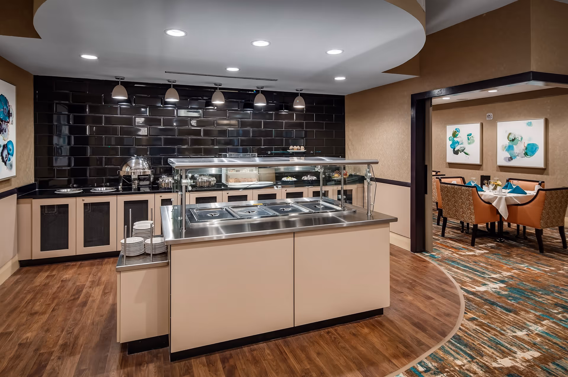 Interior view of a dining area in a senior living facility featuring a buffet station with covered food trays and plates, a black tiled backsplash, pendant lights, and a separate dining room with a table set with blue napkins and chairs.