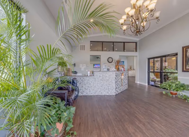Bright and spacious reception area of Regency Park Assisted Living & Memory Care with a tiled front desk, large green plants, wooden flooring, a chandelier, and glass doors leading to another room.