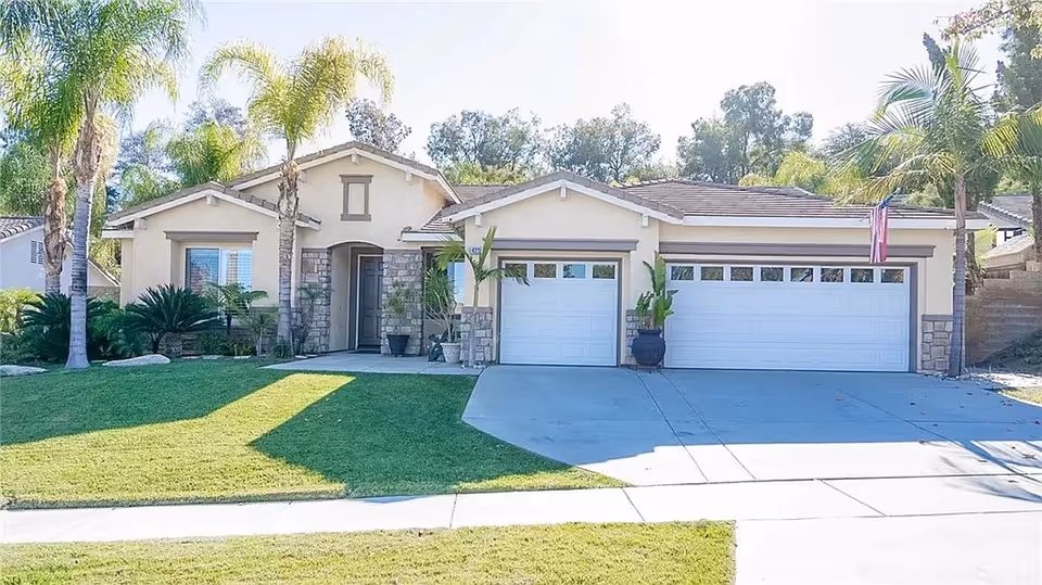 Front exterior view of a single-story house with a three-car garage, beige walls, stone accents, and a well-maintained lawn with palm trees and other plants.