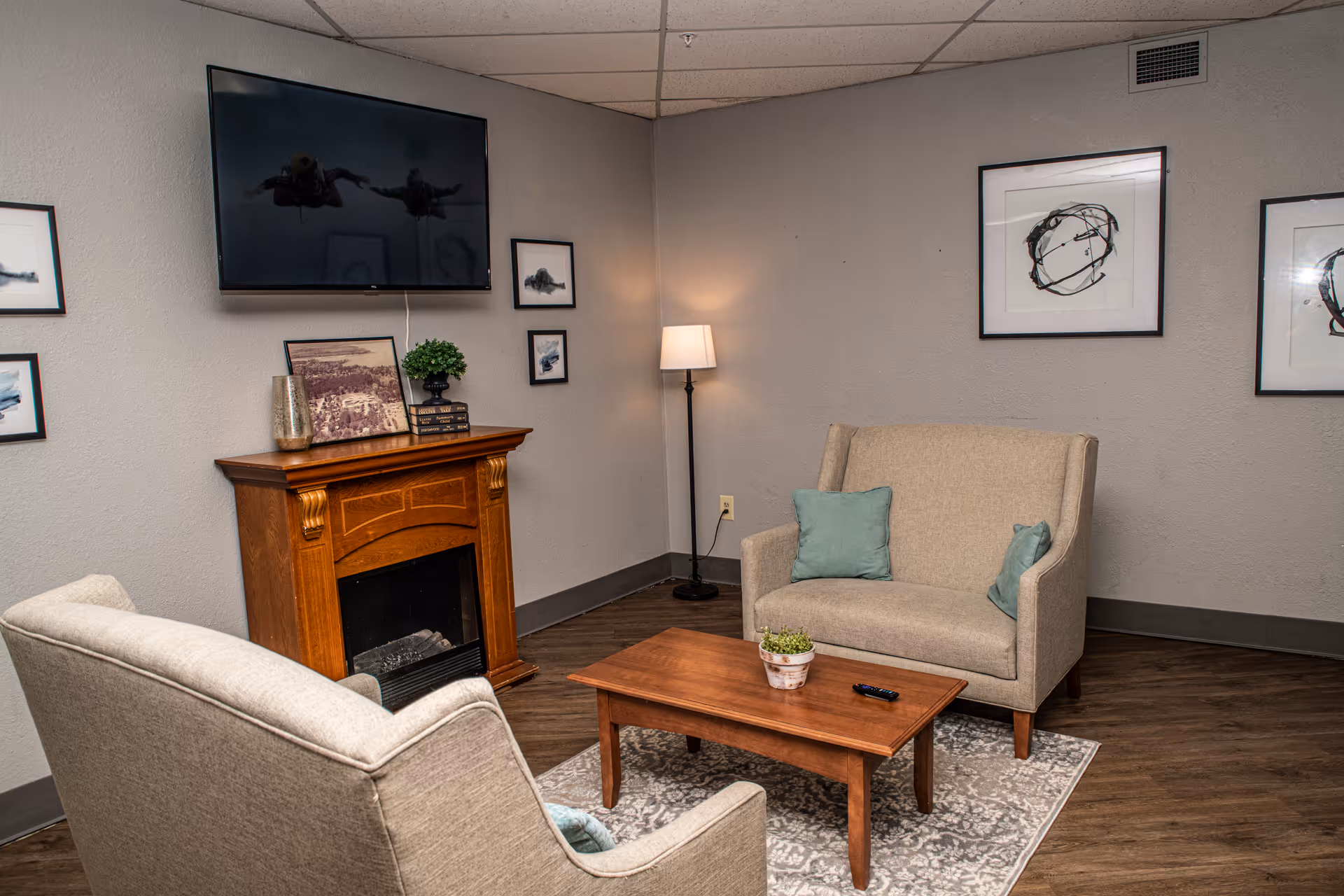 A cozy living room area with two beige upholstered armchairs facing a wooden coffee table on a patterned rug. A wooden electric fireplace mantel is against the wall with decorative items on top, and a flat-screen TV mounted above it. The walls are decorated with framed abstract art, and a floor lamp provides soft lighting in the corner.