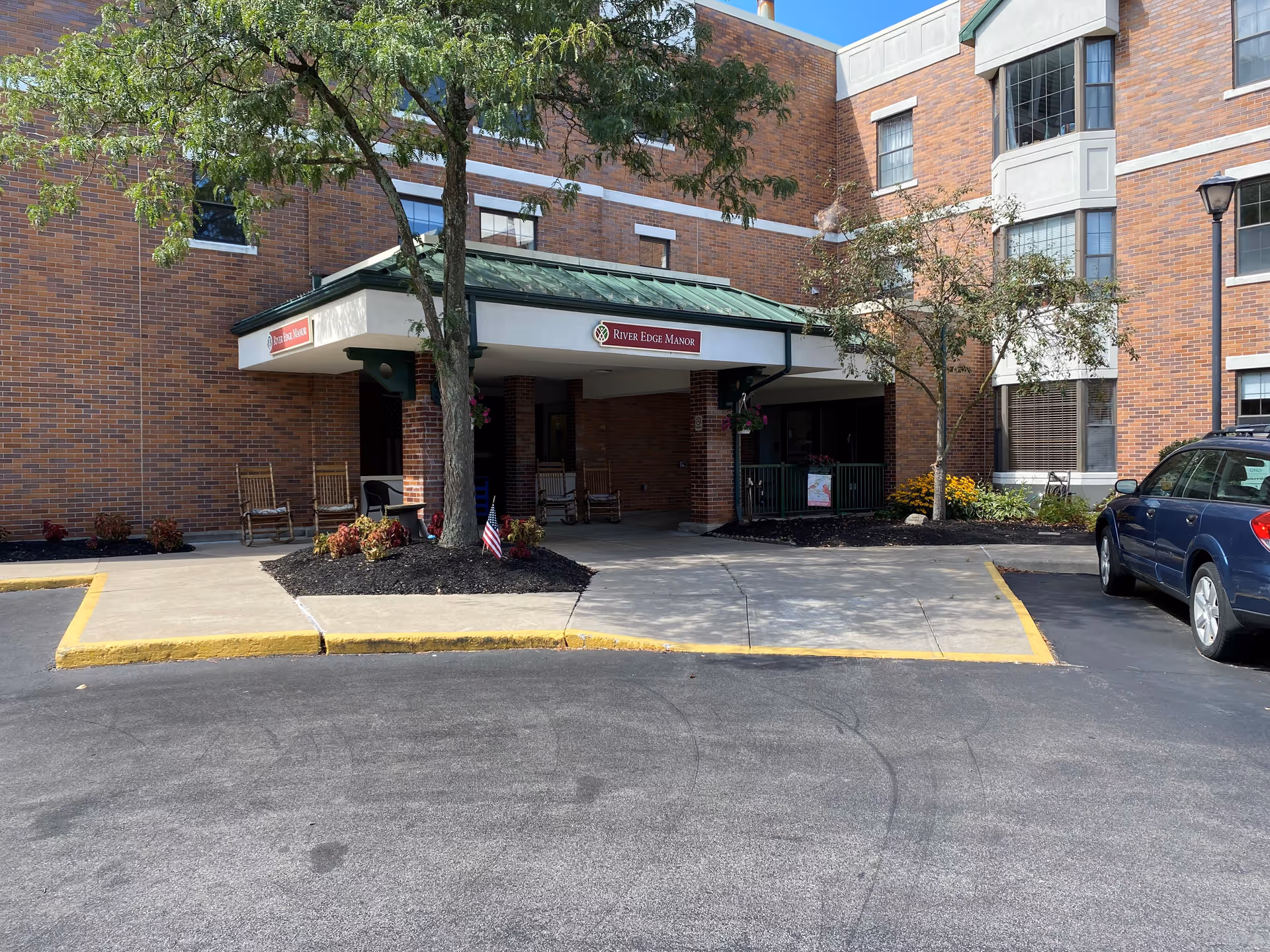 Entrance of Episcopal SeniorLife - River Edge Manor, showing a brick building with a covered entryway, green metal roof, trees, and a few chairs on the porch. A blue car is parked to the right, and there are small landscaped areas with flowers and mulch near the entrance.