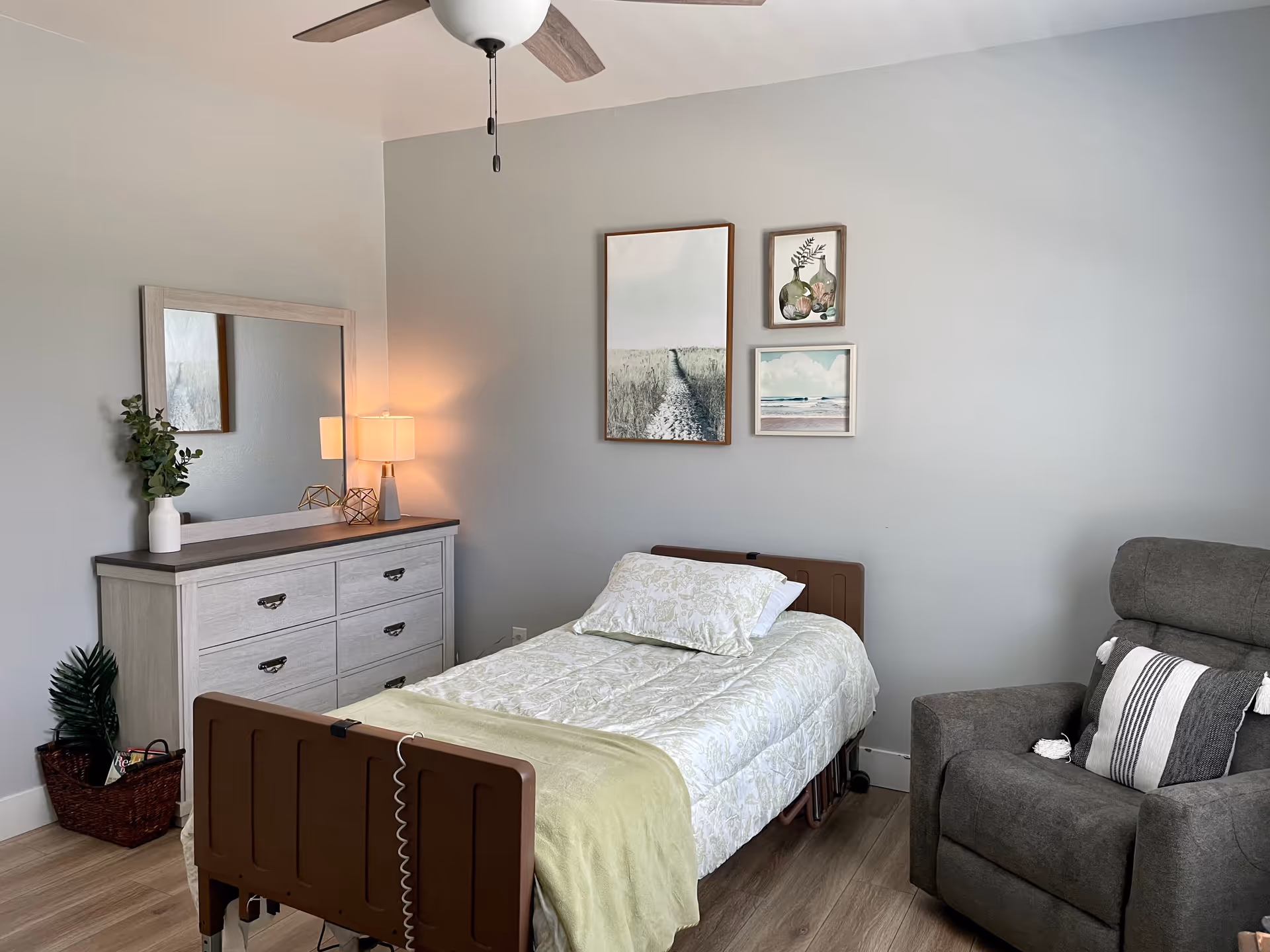 A bright bedroom featuring a single adjustable bed with light bedding, a dresser and mirror, wall art, and a gray recliner.