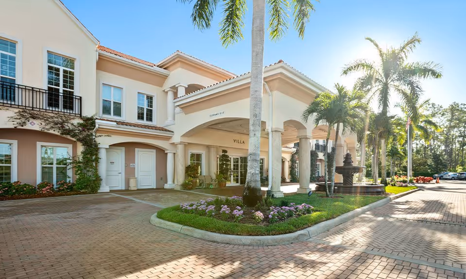 Front exterior view of Villa at Terracina Grand, showing a two-story building with a covered entrance supported by columns, palm trees, a fountain, and a paved driveway with landscaped flower beds.