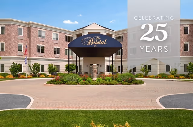 Front exterior view of The Bristal Assisted Living at Armonk building with a circular driveway, landscaped garden, and a blue canopy entrance. A banner on the right side reads 'Celebrating 25 Years'.