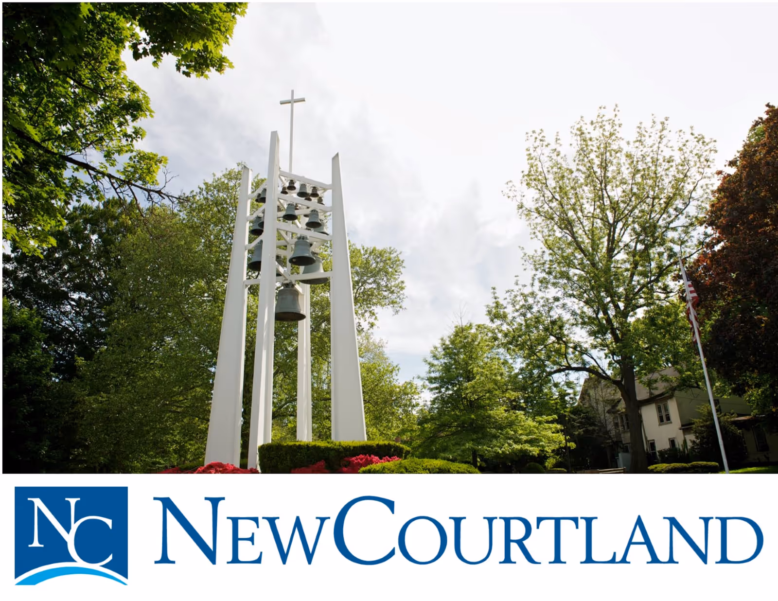 Outdoor scene featuring a tall white bell tower with multiple bells and a cross on top, surrounded by green trees and bushes under a partly cloudy sky.