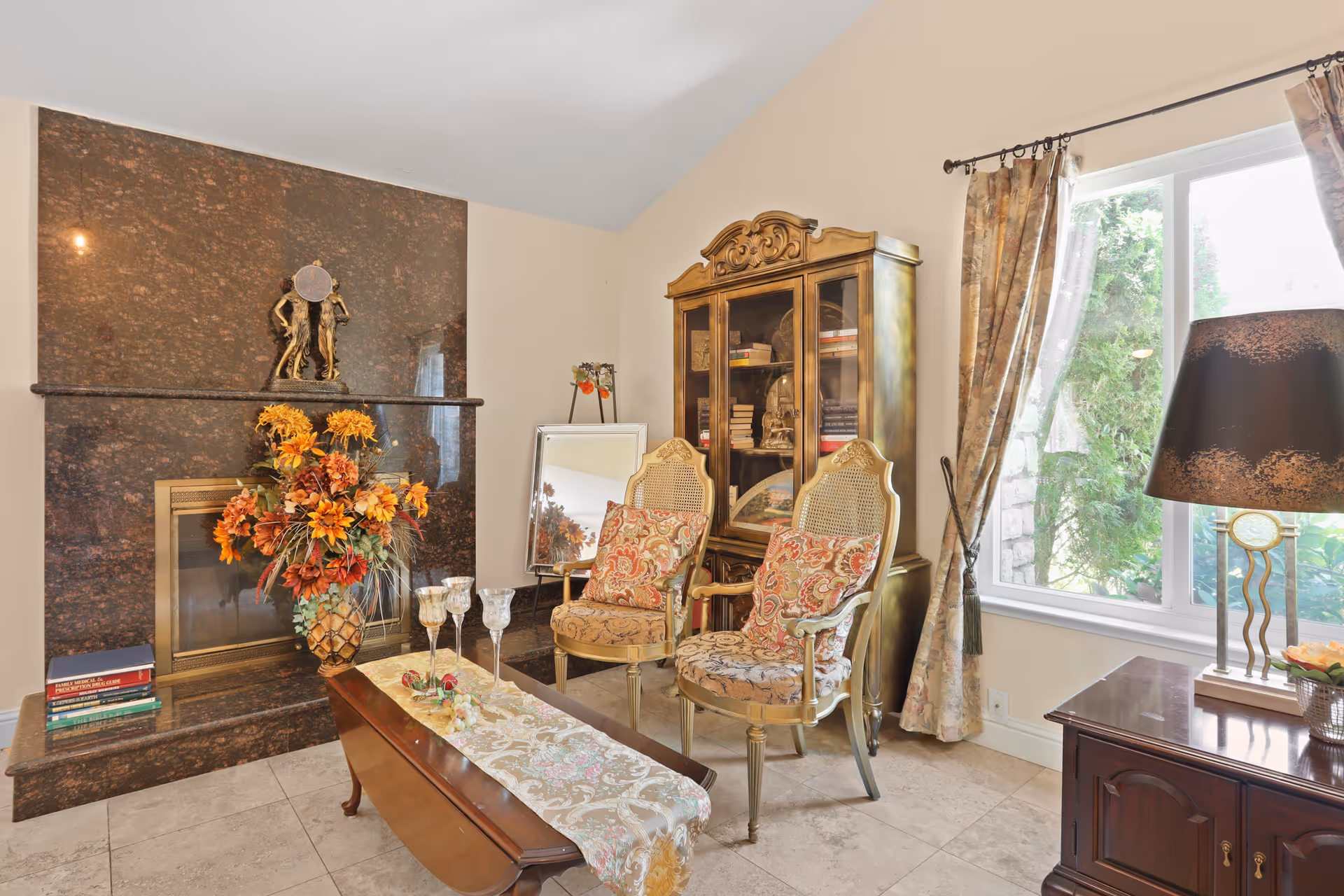 Sunlit living room with ornate upholstered chairs, a glass-front cabinet, a marble fireplace topped with a floral arrangement, and a coffee table near a window with drapes.
