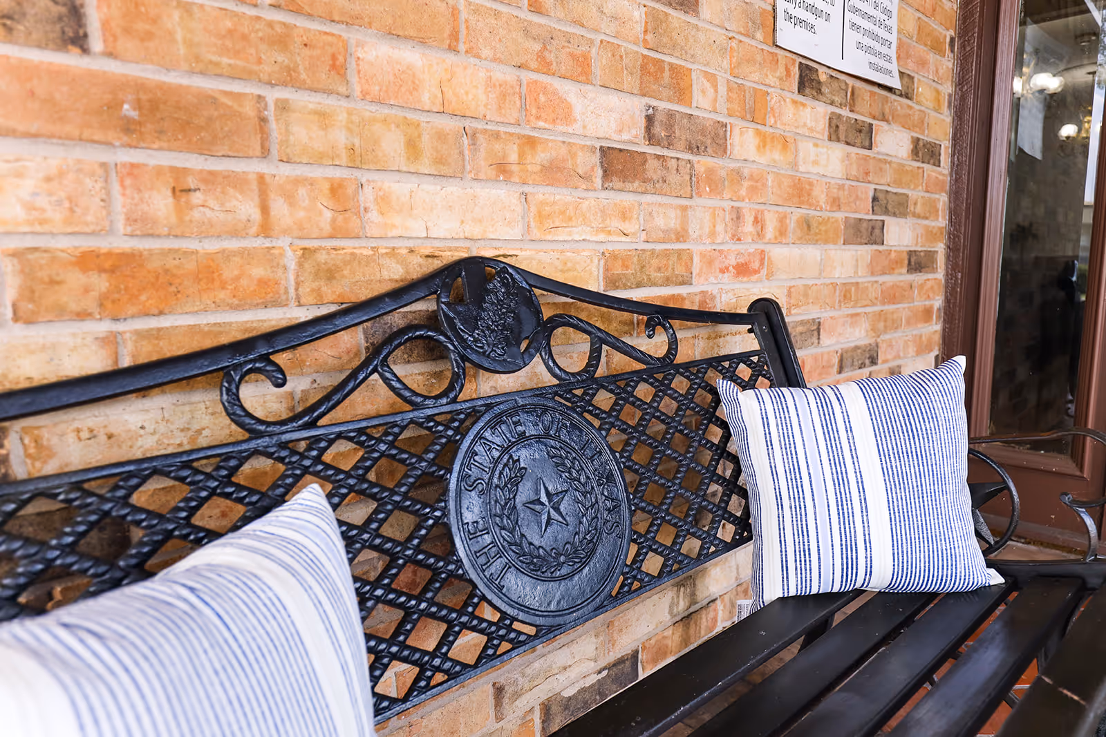 A black metal bench with decorative backrest featuring the seal of the State of Texas, placed against a brick wall. The bench has two white cushions with blue stripes. A window with a brown frame is visible on the right side.