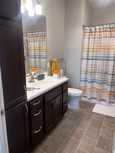 A clean bathroom featuring dark wood cabinets with silver handles, a white countertop with a sink, a large mirror above the sink, and a light fixture with three lights. There is a toilet next to the cabinets and a shower with a colorful striped curtain in shades of orange, gray, and white. A yellow towel hangs on a towel rack beside the toilet, and the floor is covered with gray tiles.