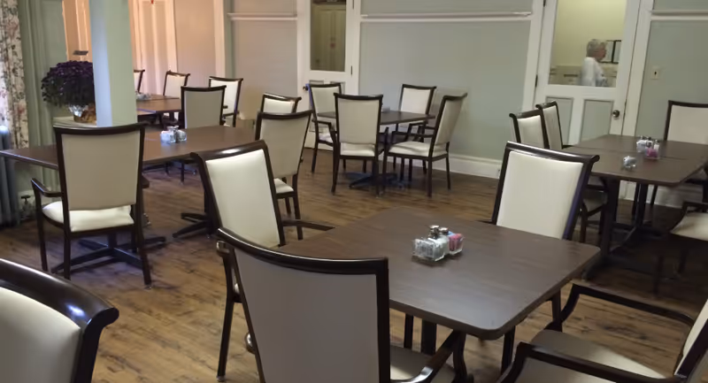 Dining room with several wooden tables and cream-upholstered chairs on hardwood flooring.