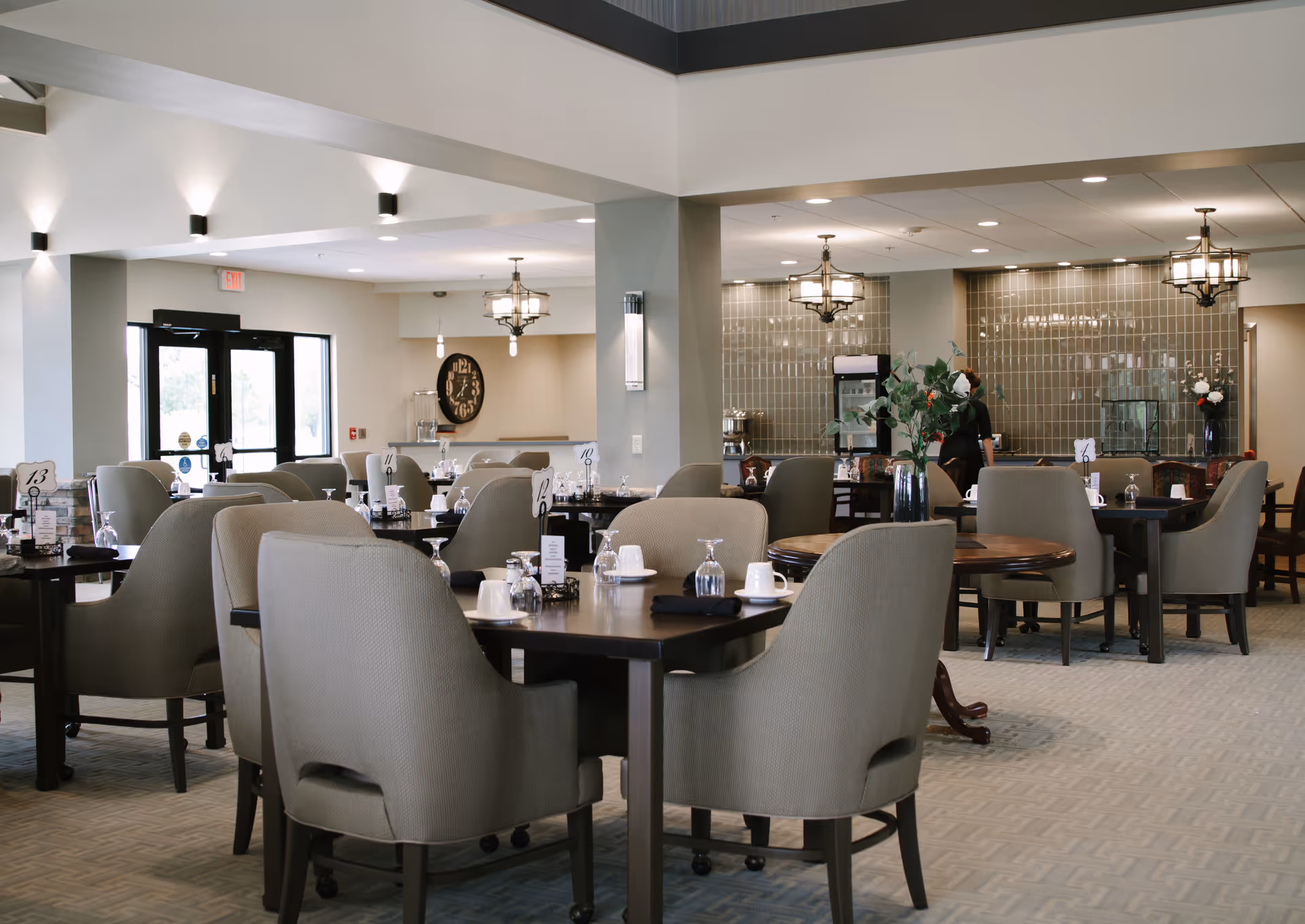 A spacious dining room in a senior living facility with multiple tables and cushioned chairs arranged neatly. Each table is set with cups, glasses, and napkins. The room features modern lighting fixtures, a large clock on the wall, and a beverage station in the background. The space is well-lit with natural light coming through glass doors.