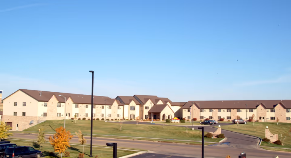 Exterior front view of a large two-story senior living building with a lawn, driveway and parked cars under a clear blue sky.