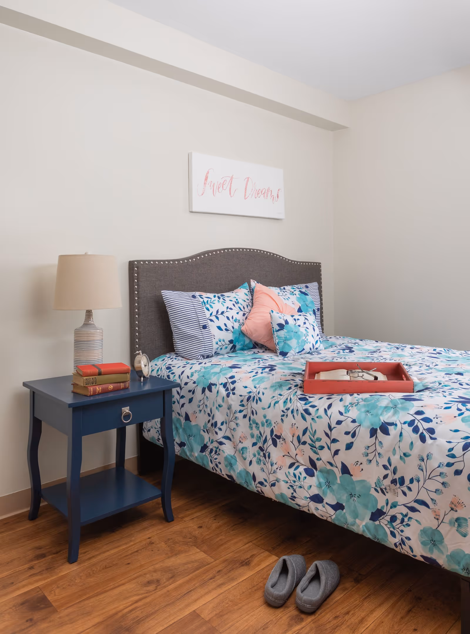 A bedroom with a bed featuring a gray upholstered headboard and floral bedding in shades of blue and teal. There are several pillows including a pink one. A red tray with an open book and glasses rests on the bed. Next to the bed is a blue nightstand with a lamp, three stacked books, and a small clock. A pair of gray slippers is on the wooden floor beside the bed. A wall decoration above the bed reads 'Sweet Dreams' in pink script.