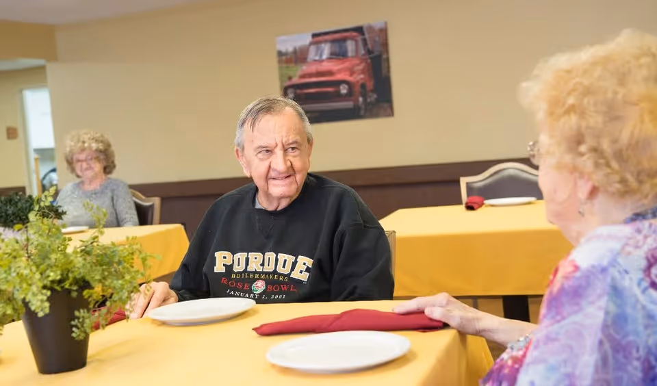Two elderly people sitting at a table with yellow tablecloths in a dining area. One man wearing a black Purdue sweatshirt is looking at a woman with curly hair and glasses. There is a potted plant on the table and a picture of a red truck on the wall in the background.