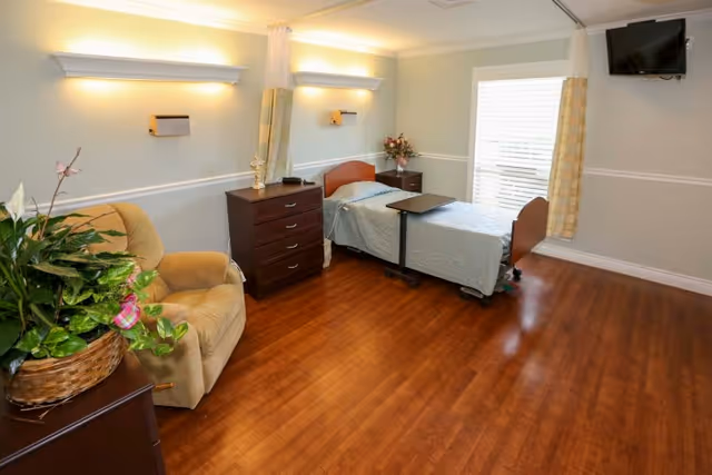 A well-lit senior living bedroom with a single bed covered in a light blue bedspread, a dark wooden chest of drawers, a beige recliner chair, a small table on wheels next to the bed, a wall-mounted TV, and a window with light-colored curtains. There is a potted plant on a small table near the chair and wooden flooring throughout the room.
