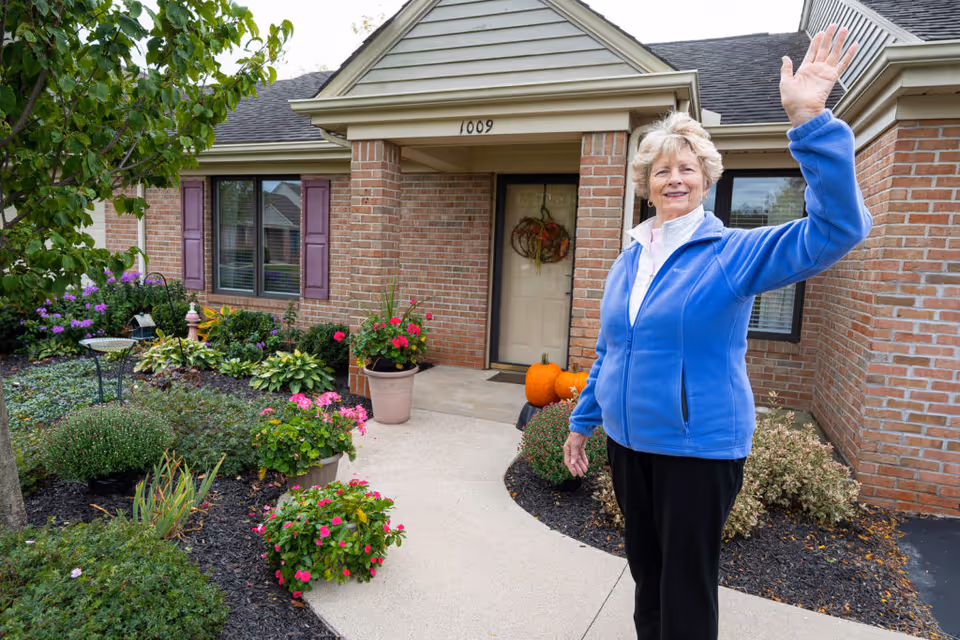 An elderly woman wearing a blue jacket waves while standing on a curved walkway leading to the front door of a brick house with purple shutters. The garden around the walkway is well-maintained with various green plants and colorful flowers. There are pumpkins placed near the front door, suggesting a fall season.