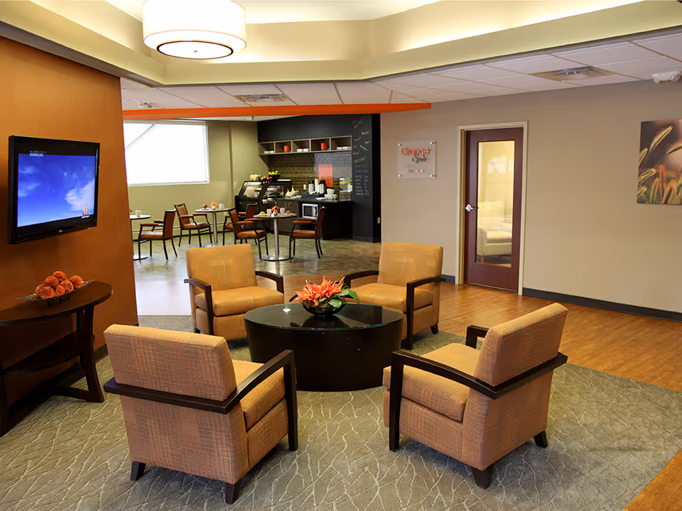 A cozy seating area with four cushioned armchairs arranged around a circular black coffee table with a flower centerpiece. In the background, there is a small dining area with tables and chairs, a TV mounted on a brown wall, and a kitchenette area labeled 'Orange Spot' with various appliances and a chalkboard menu.