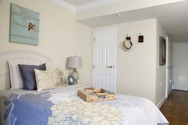 A neatly made bed with a floral patterned quilt and multiple pillows in a bright bedroom. A bedside table holds a lamp, a small vase with flowers, and a clock. The room has light-colored walls with a painting of two birds above the bed, a white door with two hooks holding a hat and a small item, and a wooden floor.