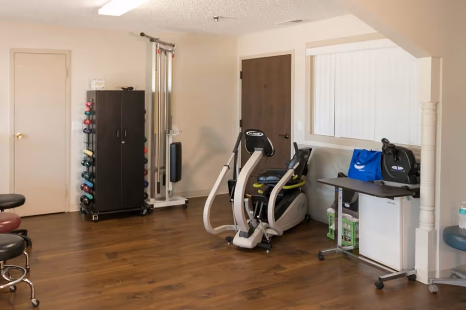 A small exercise room with wooden flooring featuring a recumbent exercise bike, a weight rack with colorful dumbbells, a cable machine, a rolling stool, and a small table with a blue bag on it. The room has beige walls, a closed door, and a window with vertical blinds.