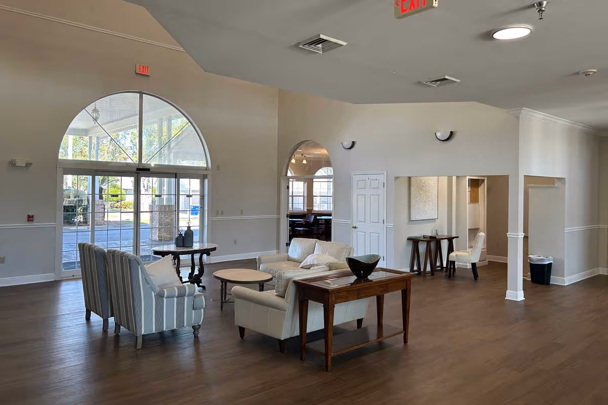 A spacious and well-lit living room area in a senior living facility with wooden flooring, a white leather sofa, two striped armchairs, a wooden coffee table, and a side table with a decorative bowl. Large arched windows and glass doors allow natural light to enter, and there is an exit sign above the door. The walls are painted light beige with white trim, and there is a small desk and chair in an adjacent room visible through an open doorway.