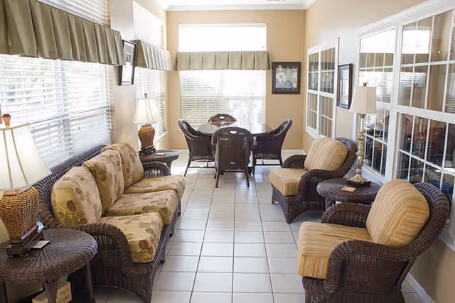 A bright, cozy sitting area with wicker furniture including a floral-patterned sofa, two striped armchairs, and a small round table with a lamp. Large windows with blinds and valances let in natural light, and a small dining table with four wicker chairs is visible at the far end of the room.