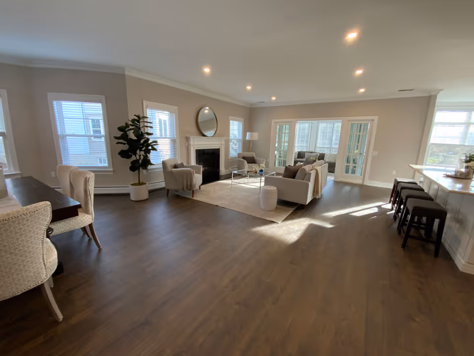 A spacious and well-lit living room area with hardwood floors, neutral-colored walls, and multiple windows allowing natural light. The room features a fireplace with a round mirror above it, two armchairs, a sofa, a coffee table, a floor lamp, and a potted plant. To the right, there is a kitchen counter with bar stools, and to the left, a dining table with upholstered chairs. French doors lead to another room with additional seating.