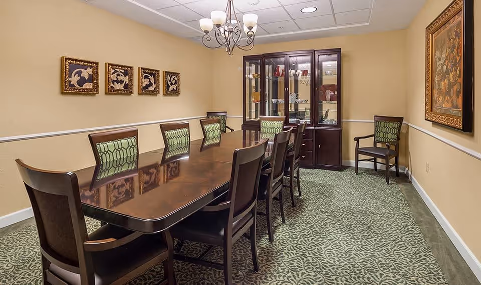 A formal dining room with a long polished wooden table surrounded by ten chairs with green patterned upholstery. The room has beige walls adorned with framed artwork, a chandelier hanging from the ceiling, and a dark wooden china cabinet displaying glassware. The floor is covered with a green patterned carpet.