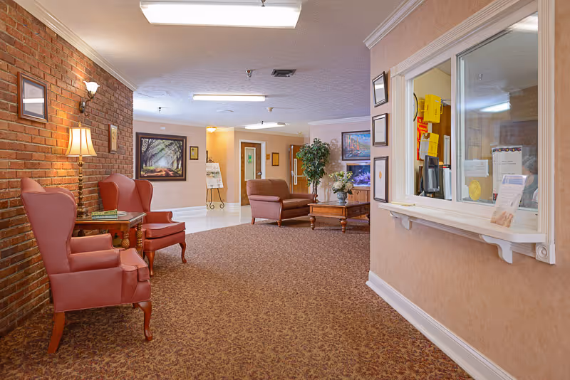 Interior view of a senior living facility lobby or common area with two red armchairs and a small table with a lamp on the left, a brick wall behind them, a carpeted floor, a reception window on the right, and a seating area with a sofa and coffee table in the background.