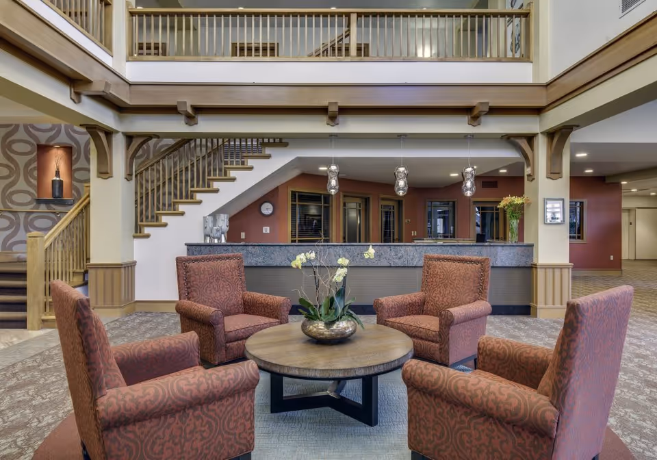 A spacious lobby area with four patterned armchairs arranged around a round wooden coffee table with a decorative plant centerpiece. Behind the seating area is a reception desk with three hanging pendant lights above it. To the left, there is a staircase leading to an upper level with wooden railings. The walls are painted in warm tones with wood accents and decorative elements.