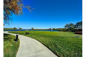 Curving paved walkway through a green lawn with benches and trees overlooking a lake under a clear blue sky.