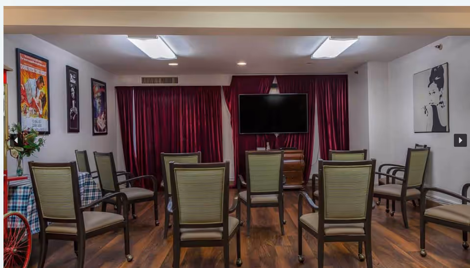 A common activity room with chairs arranged facing a wall-mounted TV and burgundy curtains, with framed art on the walls.