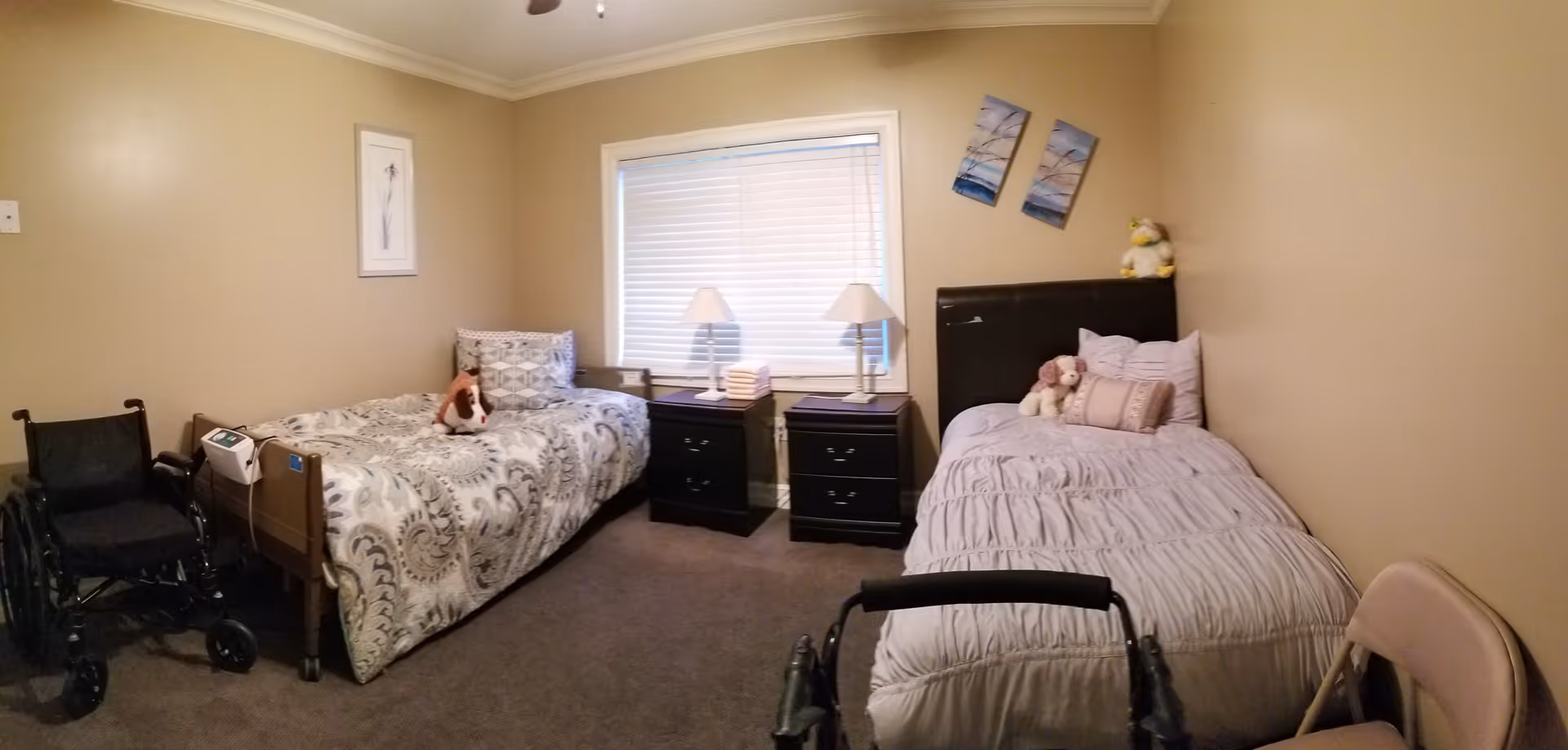 A small bedroom with two single beds separated by two black nightstands with lamps. Each bed has pillows and stuffed animals on it. There is a wheelchair next to the bed on the left and a walker in front of the bed on the right. The walls are beige with two paintings above the right bed and one painting on the left wall. A window with closed blinds is centered on the back wall.