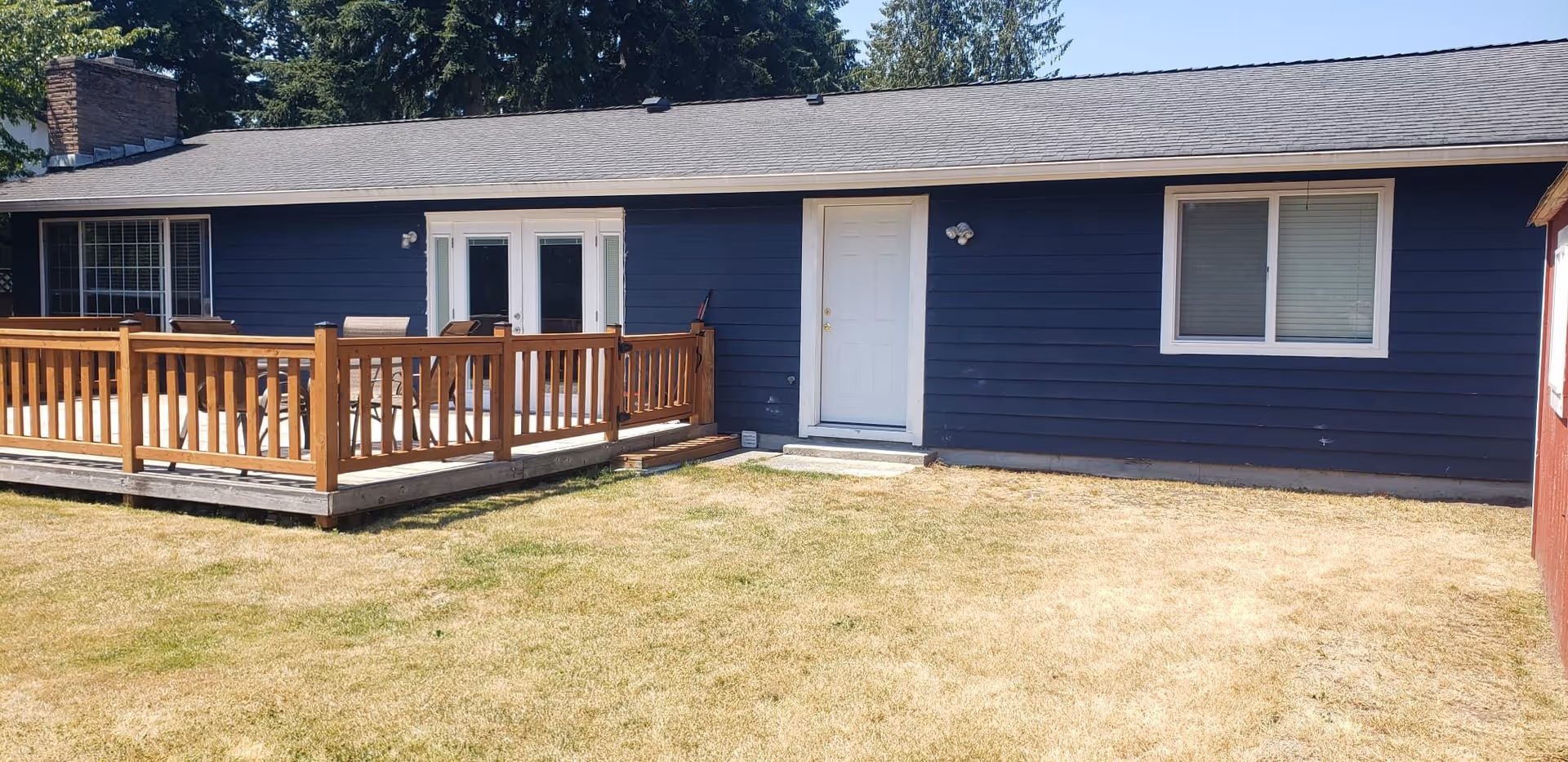 Exterior view of a single-story building with dark blue siding and a gray roof. There is a wooden deck with a railing and outdoor chairs on the left side, a white door in the center, and a window with closed blinds on the right. The building is surrounded by a dry grassy yard and some trees in the background.