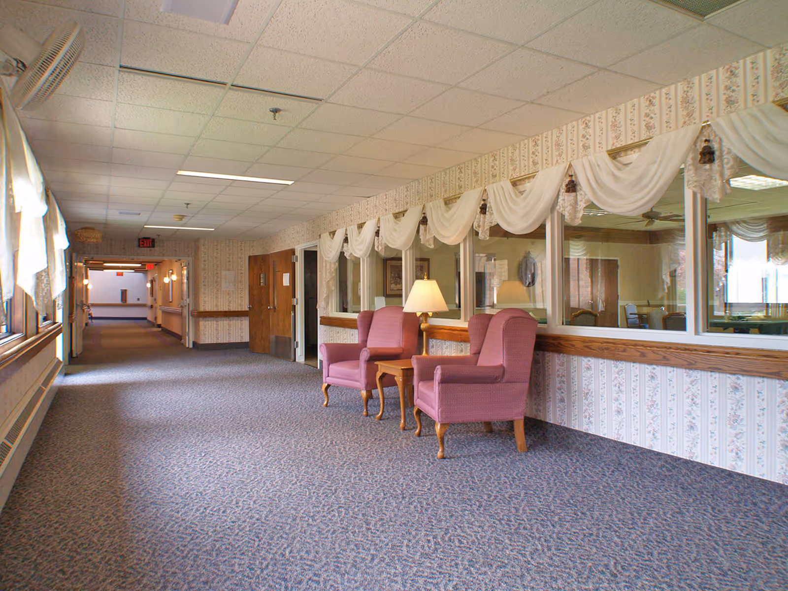 A long carpeted hallway in a senior living facility with floral wallpaper and large windows on the right side covered with white draped curtains. Two pink upholstered armchairs and a wooden side table with a lamp are placed against the wall beneath the windows. The hallway is well-lit with ceiling lights and has wooden doors and handrails along the walls.