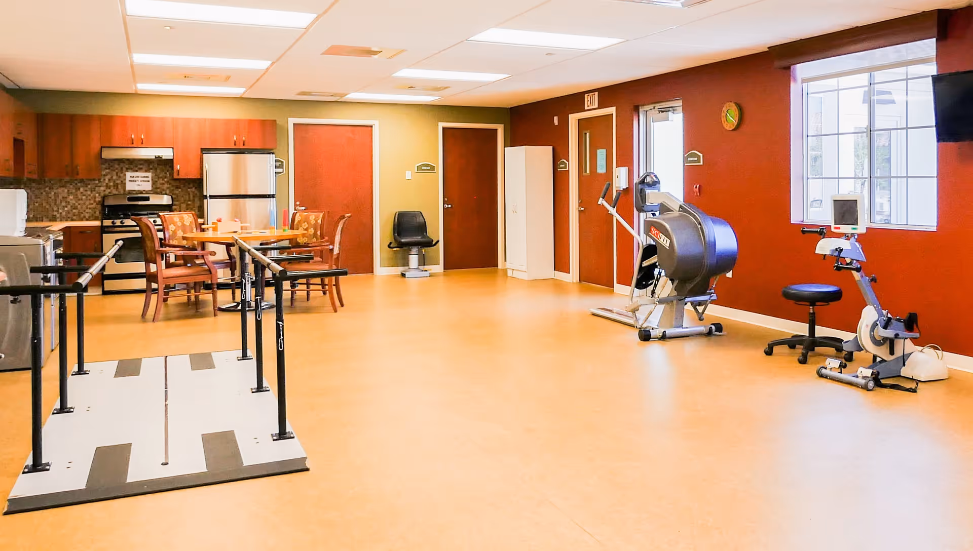 A spacious room in a senior living facility featuring a small kitchen area with a stove, refrigerator, and cabinets. There is a dining table with chairs in the middle of the room. On the right side, there are exercise equipment including a stationary bike and a rehabilitation treadmill with handrails. The walls are painted in warm tones with a large window letting in natural light.