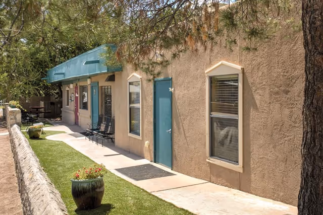 Exterior view of a single-story building with beige stucco walls and teal doors and window frames. There is a narrow concrete walkway alongside the building, bordered by a low stone wall and a strip of artificial grass with potted plants. Trees provide some shade over the area.