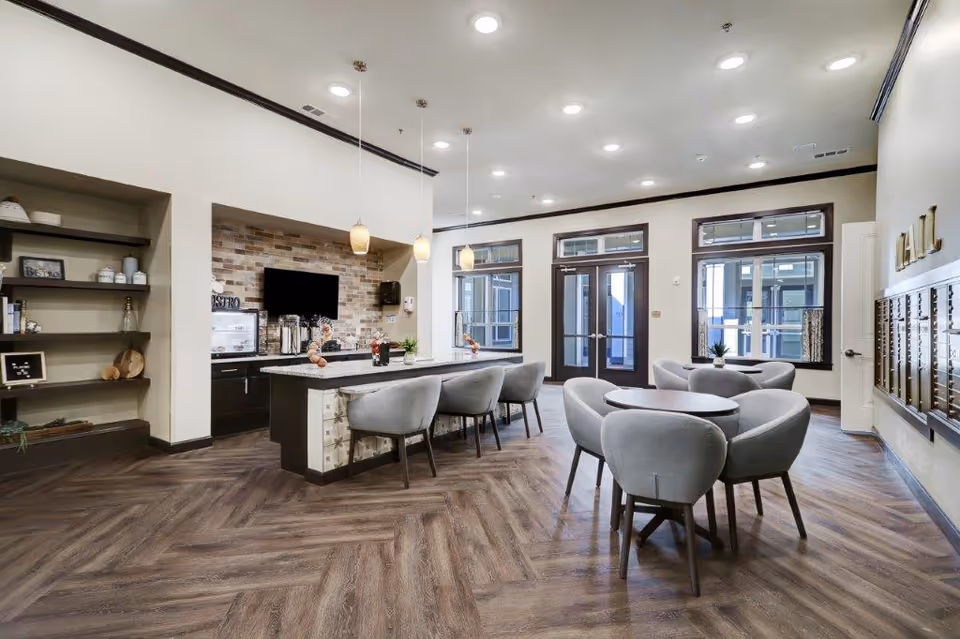 A modern common area with a kitchen island featuring four gray upholstered chairs, pendant lights hanging above, and a wall-mounted TV. There are additional round tables with gray chairs, large windows, double glass doors, and a mail area with mailboxes on the right wall. The floor has a wood pattern and the walls are light-colored with dark trim.