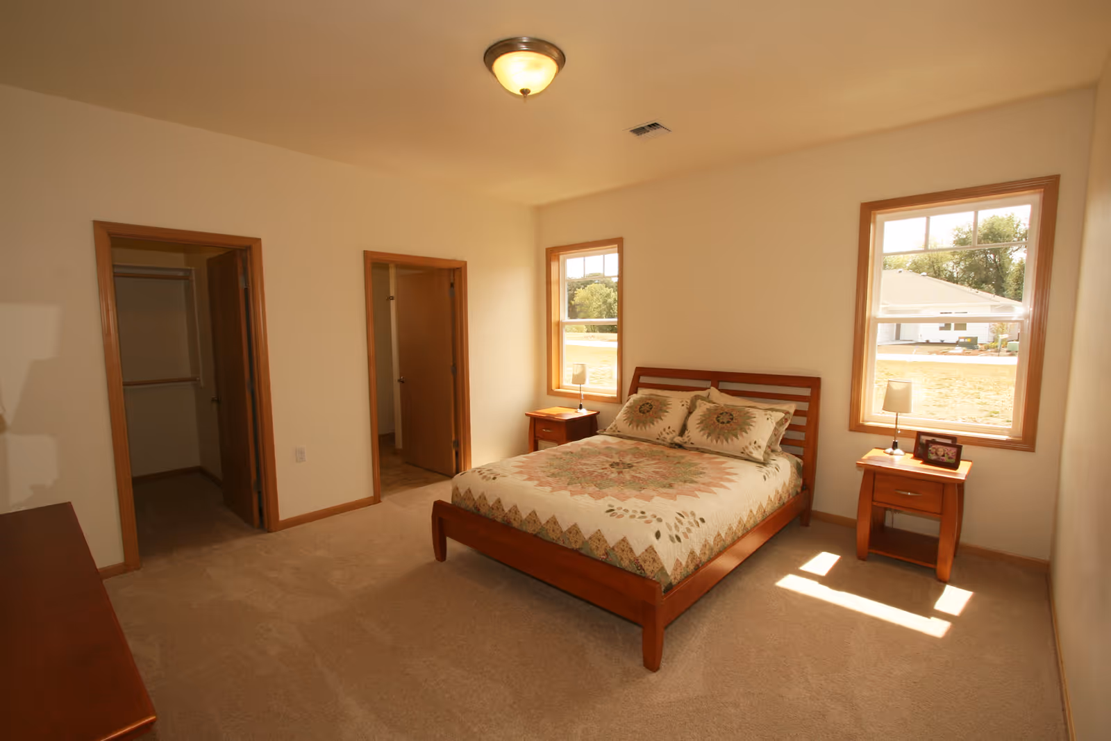 A bedroom with a wooden bed frame and a quilted bedspread featuring a floral pattern. There are two matching wooden nightstands on either side of the bed, each with a small lamp. The room has beige carpet, cream-colored walls, two windows letting in natural light, and two open doorways leading to other rooms.