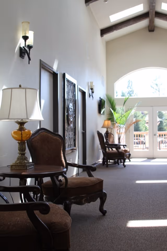 A bright hallway in a senior living facility with vintage-style armchairs and side tables along the wall. The hallway has high ceilings with exposed wooden beams, wall sconces, and large windows at the far end letting in natural light. There is a potted plant near the windows and a decorative wall piece between two doors.