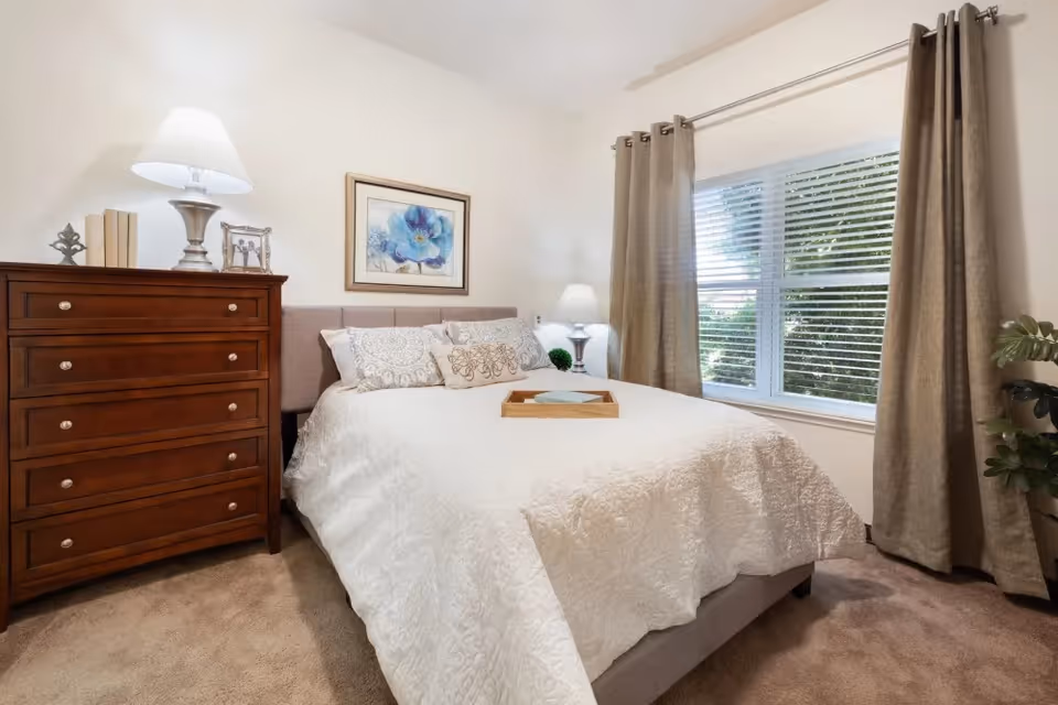 A cozy bedroom with a neatly made bed covered in a white quilt. There are decorative pillows on the bed and a wooden tray placed on top. To the left of the bed is a wooden chest of drawers with a lamp, books, and a framed photo on it. A framed floral artwork hangs on the wall above the bed. To the right, there is a window with beige curtains partially open, letting in natural light and showing greenery outside. A small potted plant is visible near the window.