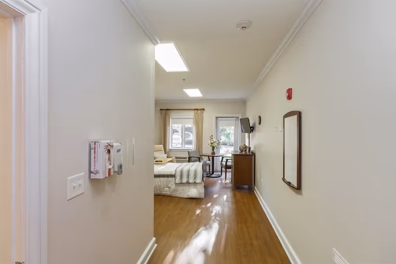 View down a hallway leading to a bedroom with a bed, a small table with two chairs, a wall-mounted TV, and a window with beige curtains. The floor is wooden, and the walls are painted light beige. There is a mirror mounted on the right wall and a fire alarm above it.