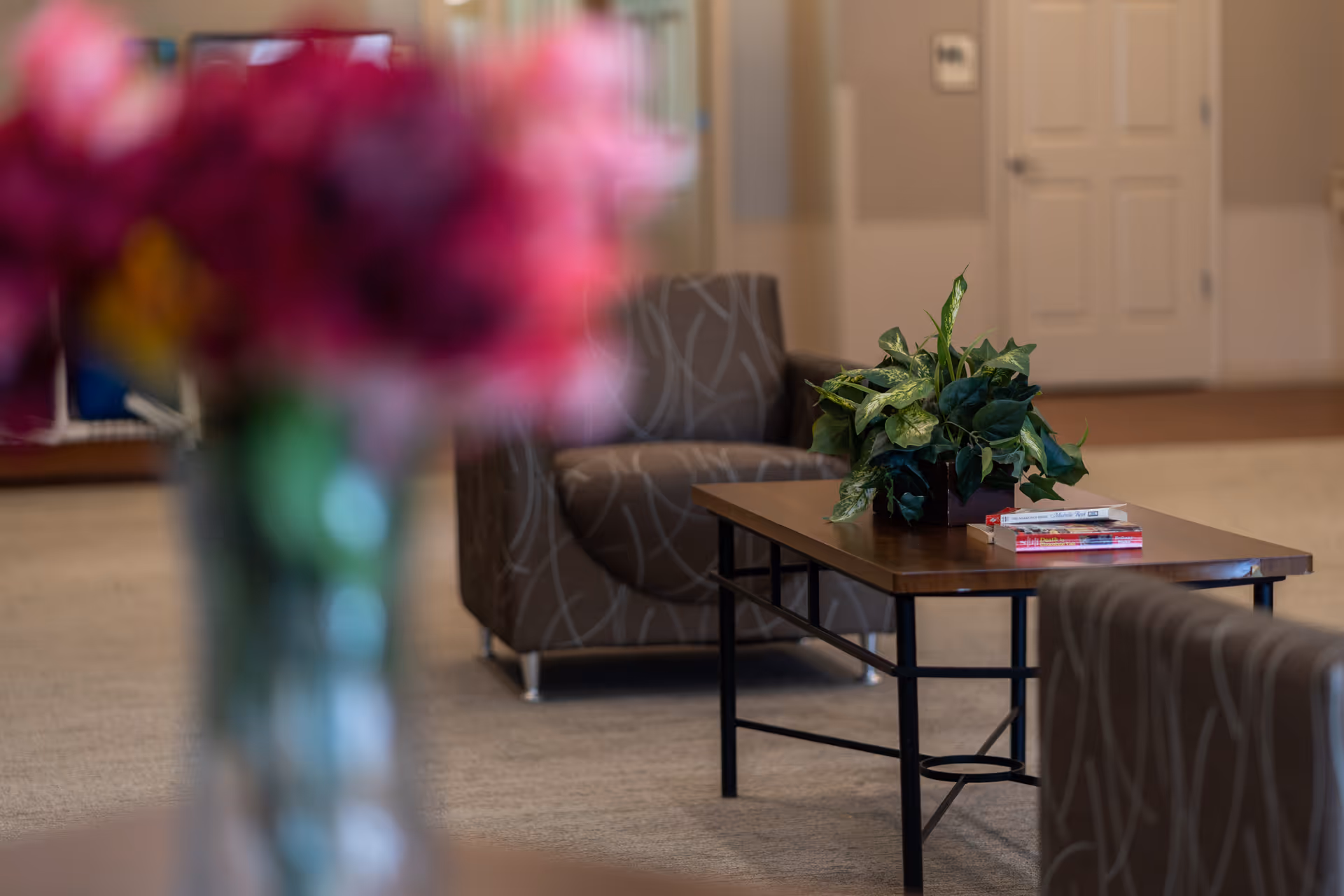 A lounge area with patterned armchairs, a coffee table holding a potted plant and books, and a blurred vase of flowers in the foreground.