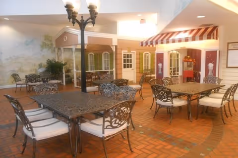 Indoor seating area with multiple metal tables and chairs with cushions on a brick floor. The space features decorative facades resembling small storefronts or houses, a vintage-style street lamp, and a popcorn machine under a striped awning.