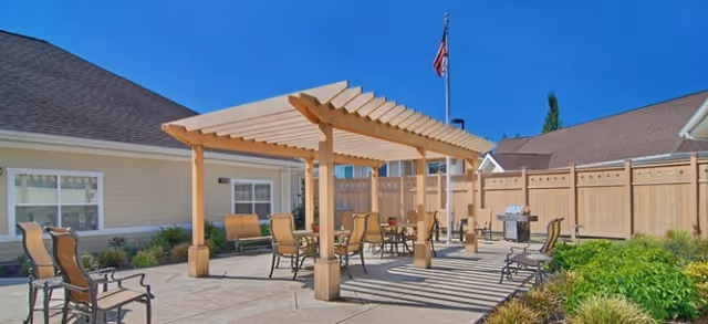 Sunlit outdoor courtyard with a wooden pergola, patio tables and chairs beside a single-story building and an American flag.