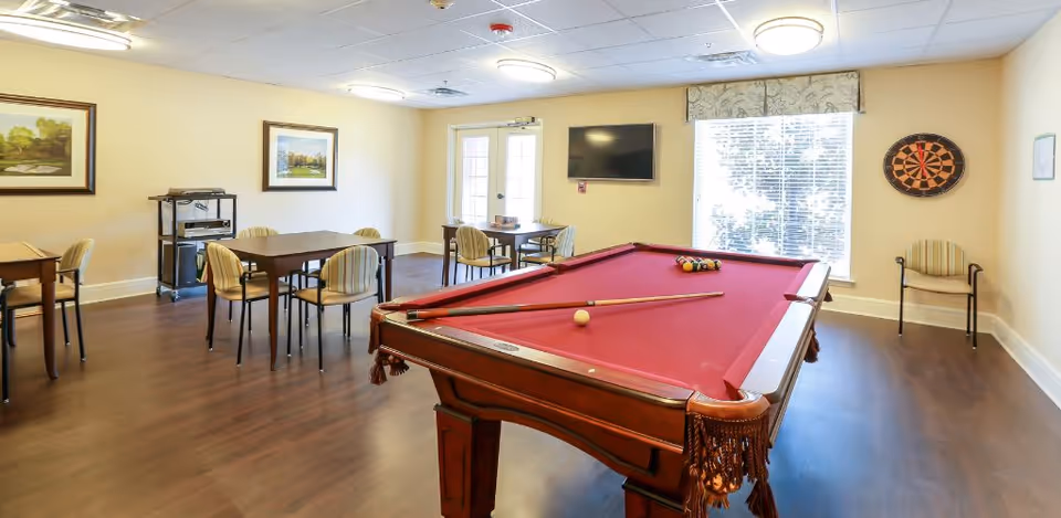 A recreational room with a red pool table in the center, several tables and chairs arranged around the room, a dartboard on the wall, a flat-screen TV mounted near a window with blinds, and framed artwork on the walls. The room has wooden flooring and soft yellow walls.