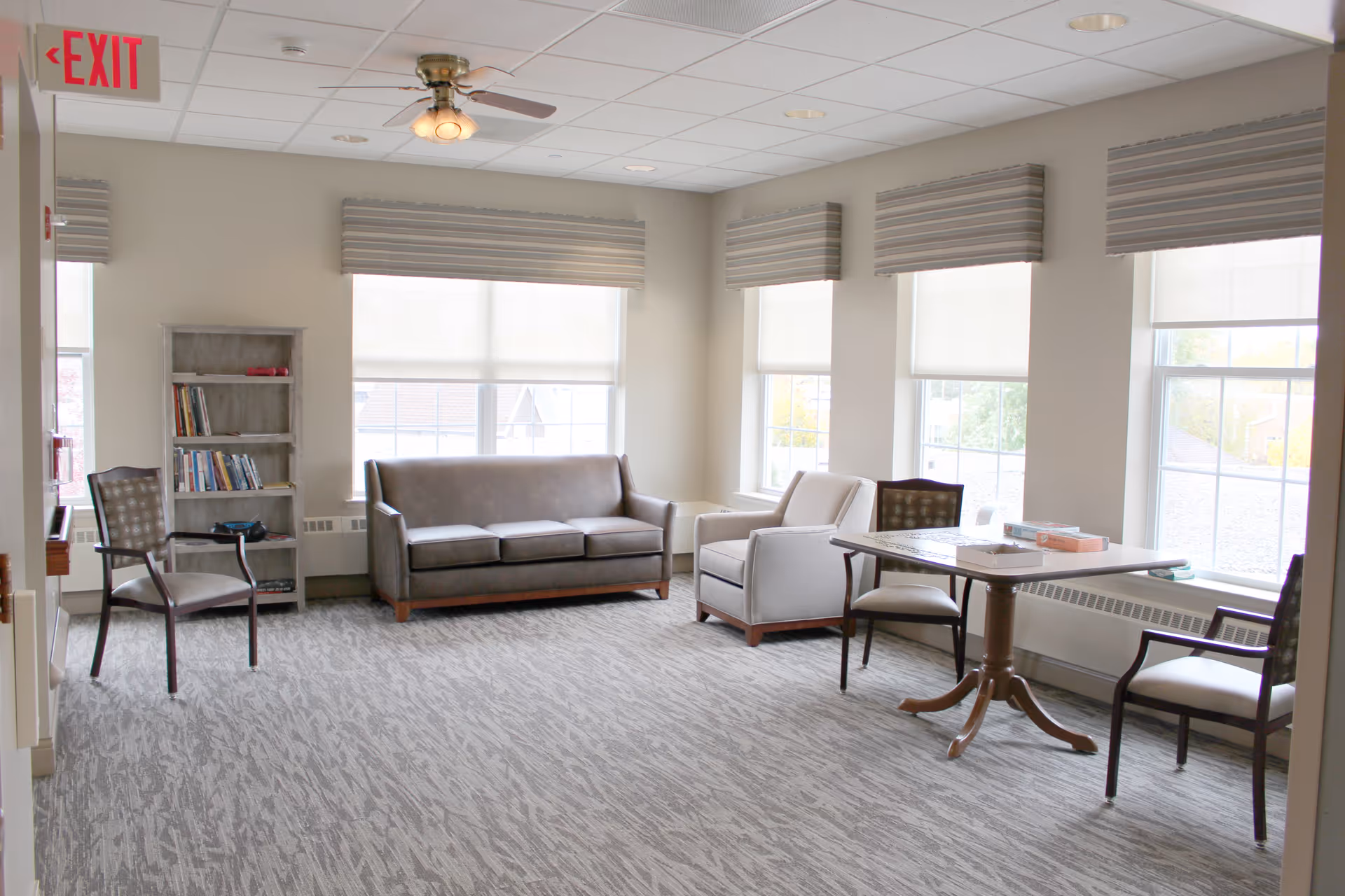 A bright and spacious living area with large windows covered by blinds. The room features a gray sofa, a light gray armchair, a wooden table with four chairs, and a small bookshelf with books. The carpet is patterned in shades of gray, and there is a ceiling fan with lights. An exit sign is visible on the left side of the image.