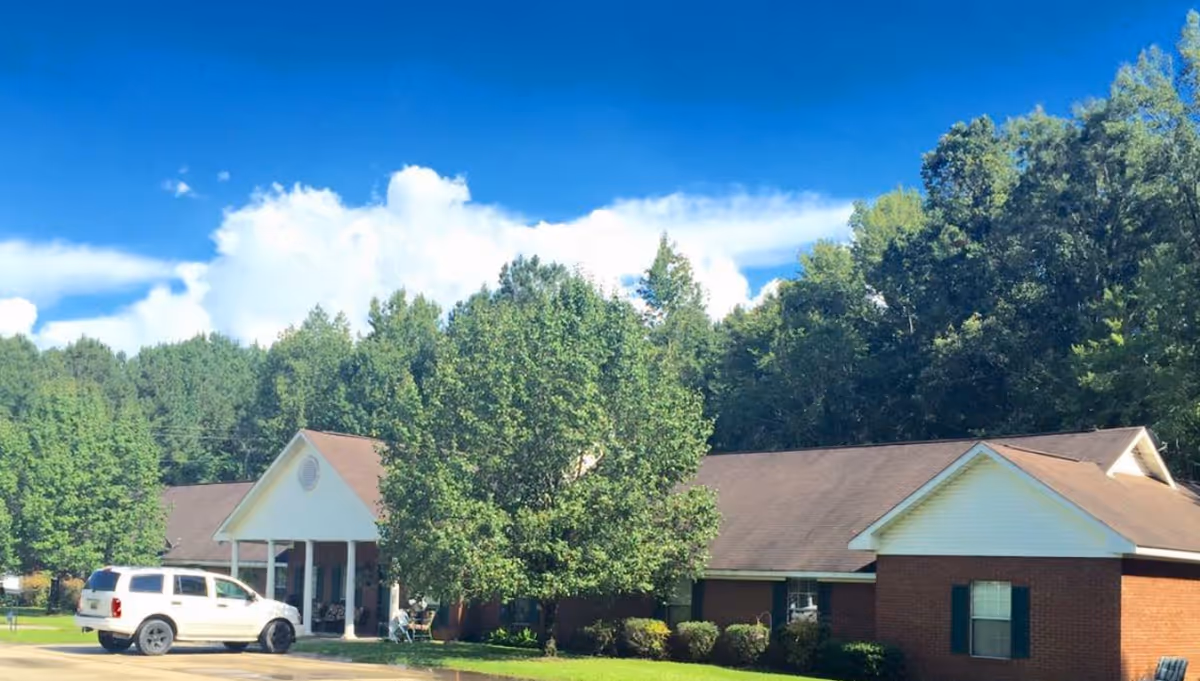 Exterior view of a single-story brick building with a brown roof, white trim, and green shutters, surrounded by trees and greenery under a blue sky with some clouds. A white SUV is parked in front of the building near the entrance with white columns.