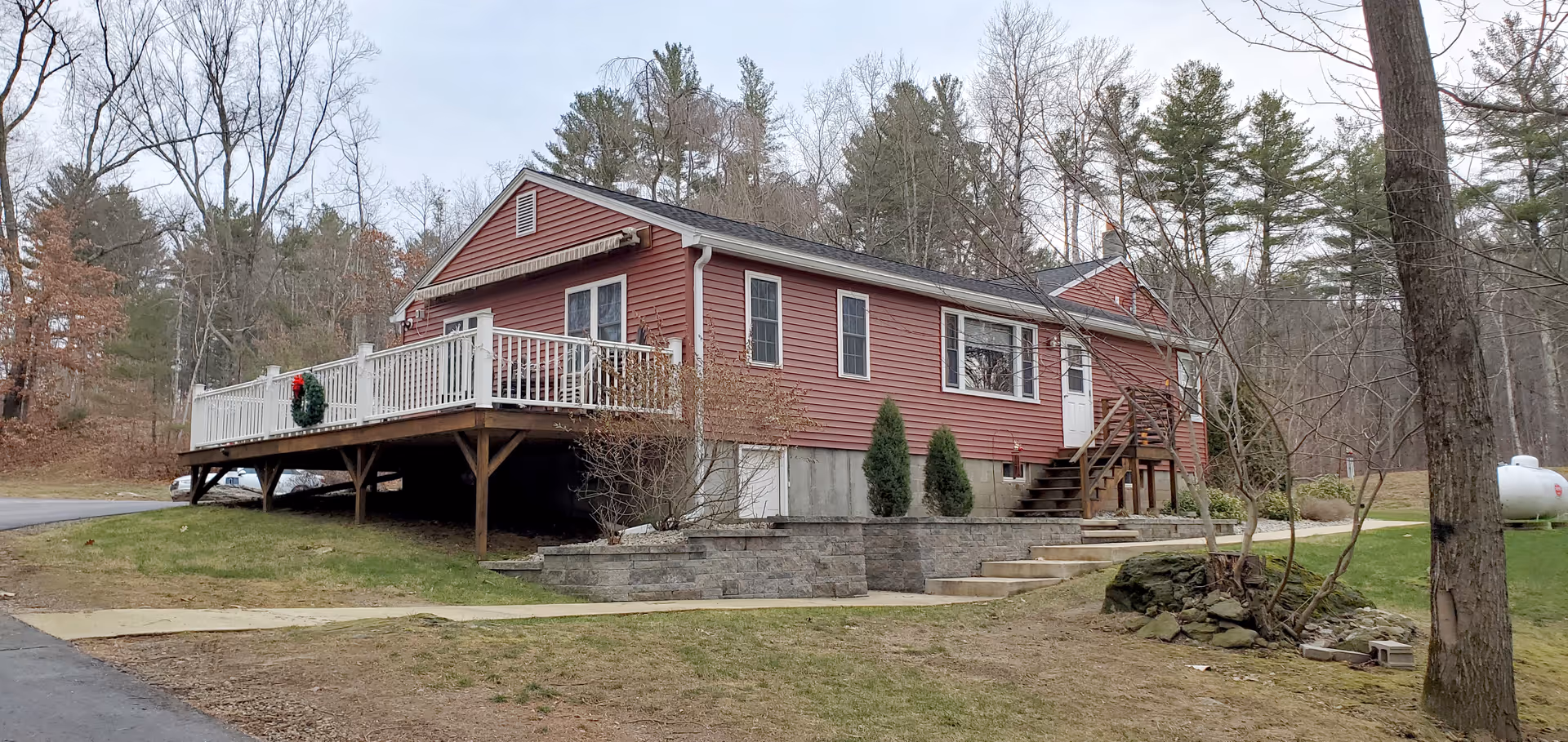 Exterior view of a single-story red house with white trim and a wooden deck decorated with a holiday wreath. The house is surrounded by grass, trees, and a paved driveway, with a wooded area in the background.